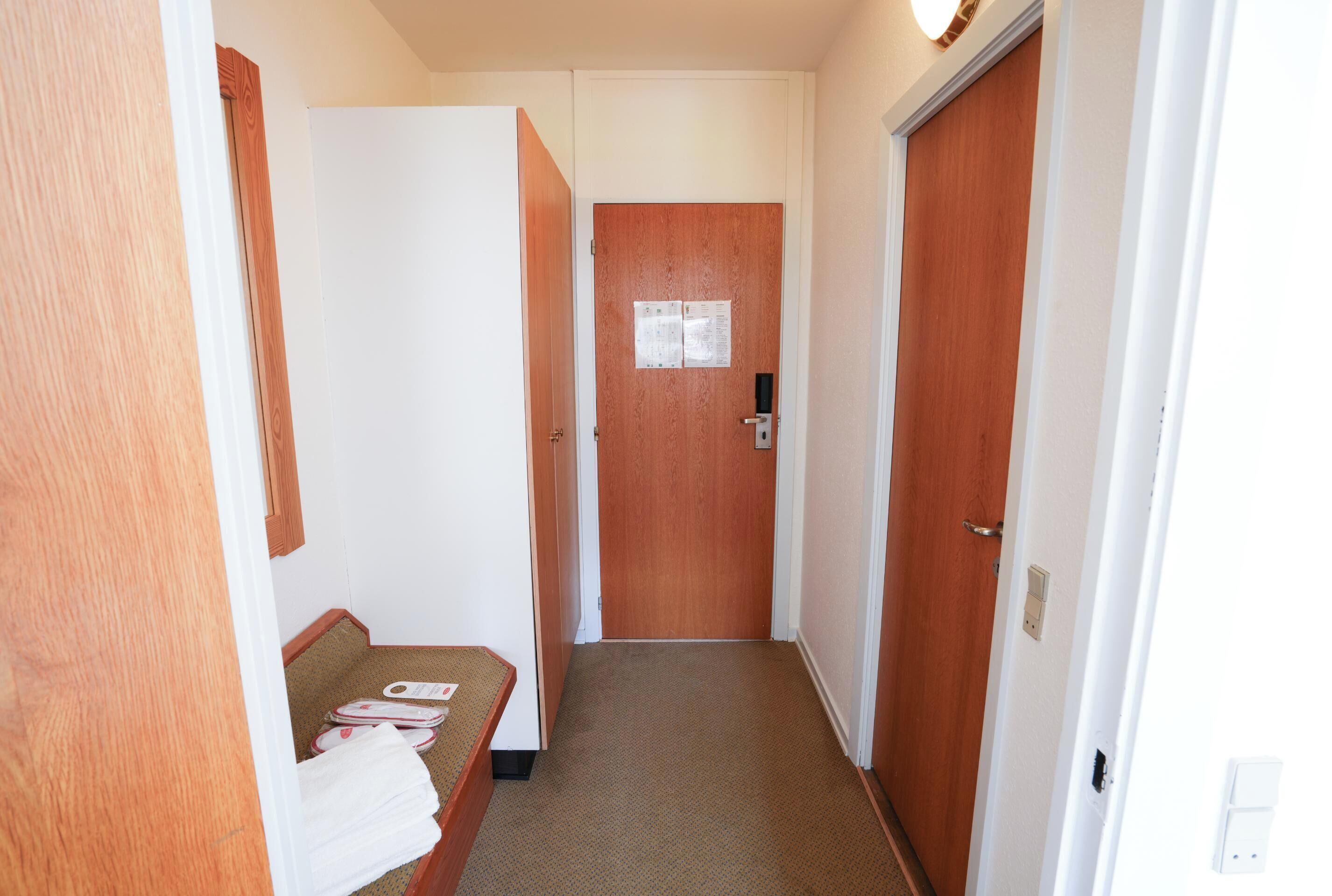 Un pasillo estrecho con una puerta de madera al final, una pared blanca y una pequeña zona de descanso con una toalla y pantuflas.