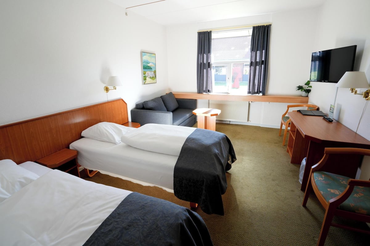 A hotel room with two single beds, a sofa, a desk with a chair, a television, and a window with curtains.