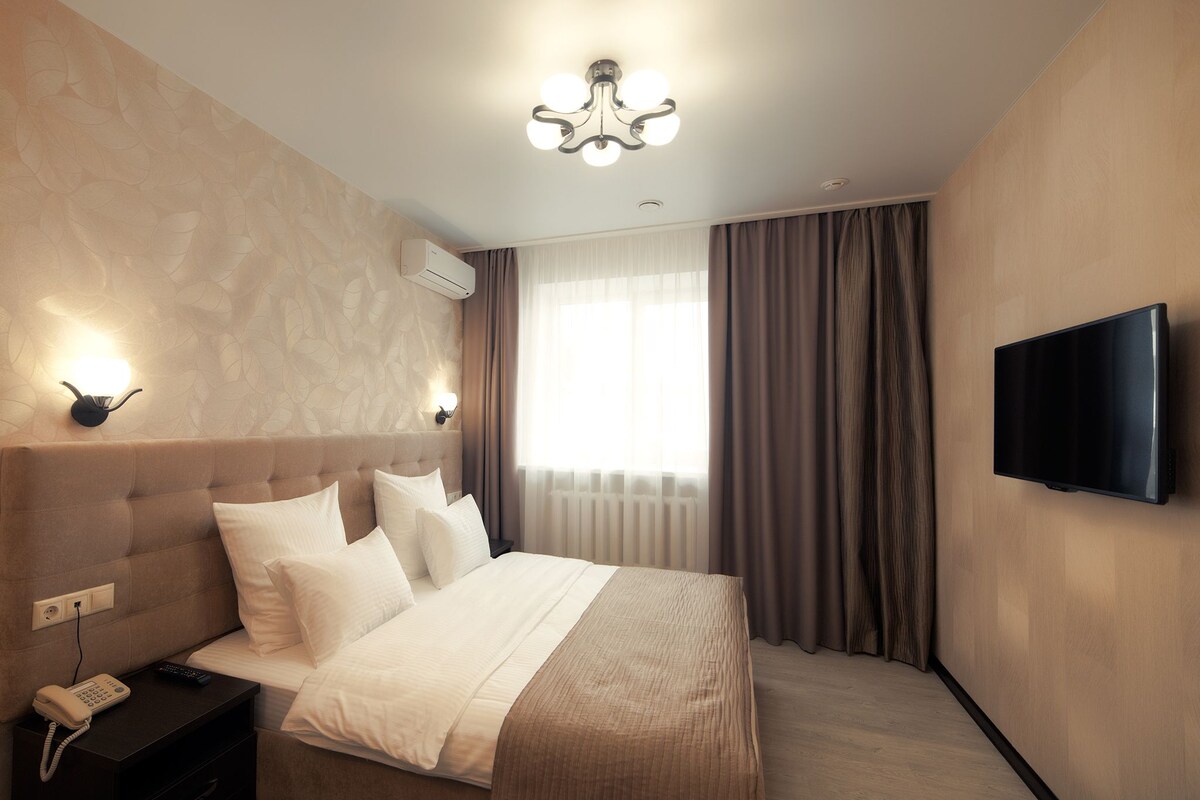 A modern hotel room with a large bed, a flat-screen TV mounted on the wall, a bedside telephone, and a window with curtains.