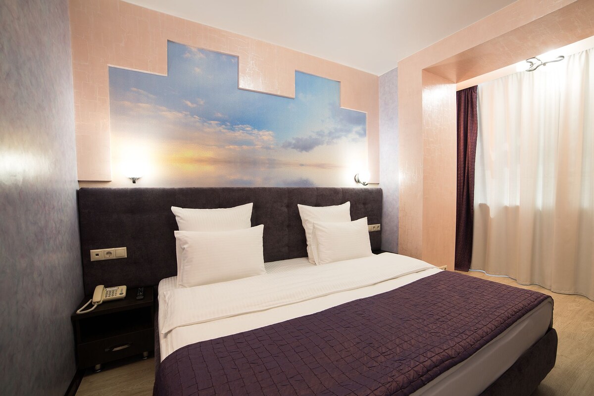 A hotel room with a large bed, a headboard with a cityscape mural, a bedside telephone, and a window with curtains.