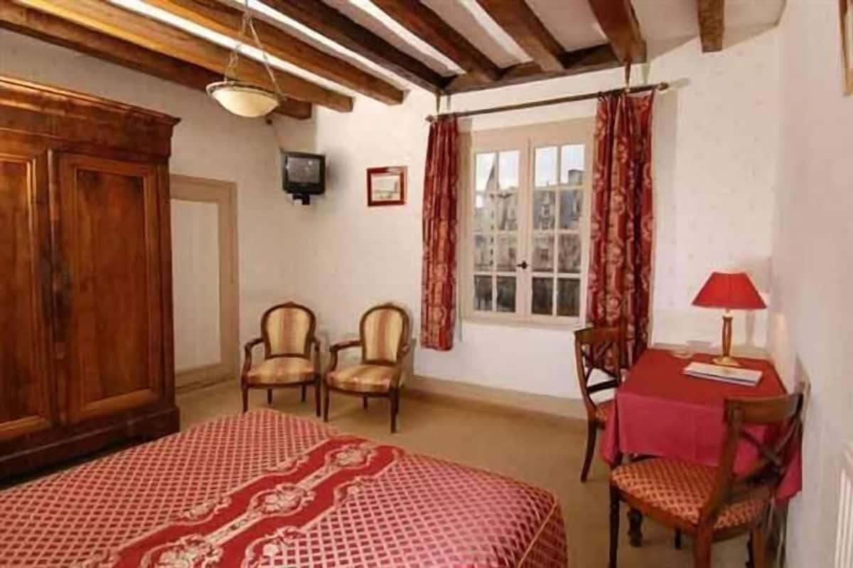 Une pièce comprenant un lit, deux chaises, une table recouverte d’une nappe rouge et une fenêtre avec des rideaux.