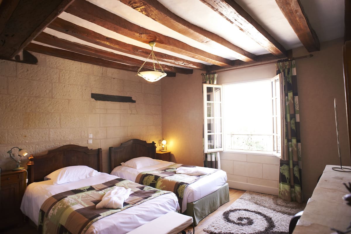 Une chambre avec deux lits, des poutres en bois, un mur en briques, une fenêtre avec des rideaux et une petite table avec une lampe.