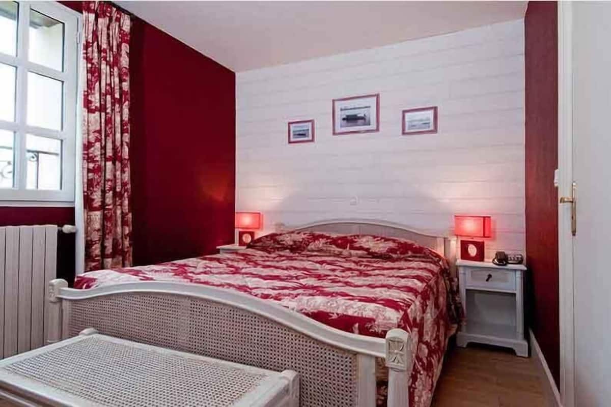 Une chambre avec un lit, deux tables de chevet, une fenêtre avec des rideaux et trois tableaux encadrés accrochés au mur.