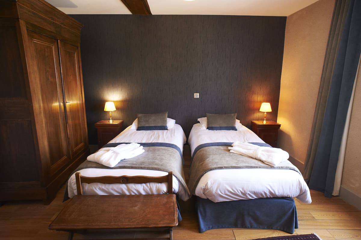 Une chambre d’hôtel avec deux lits simples, une armoire en bois, des tables de chevet avec des lampes et une petite table en bois.