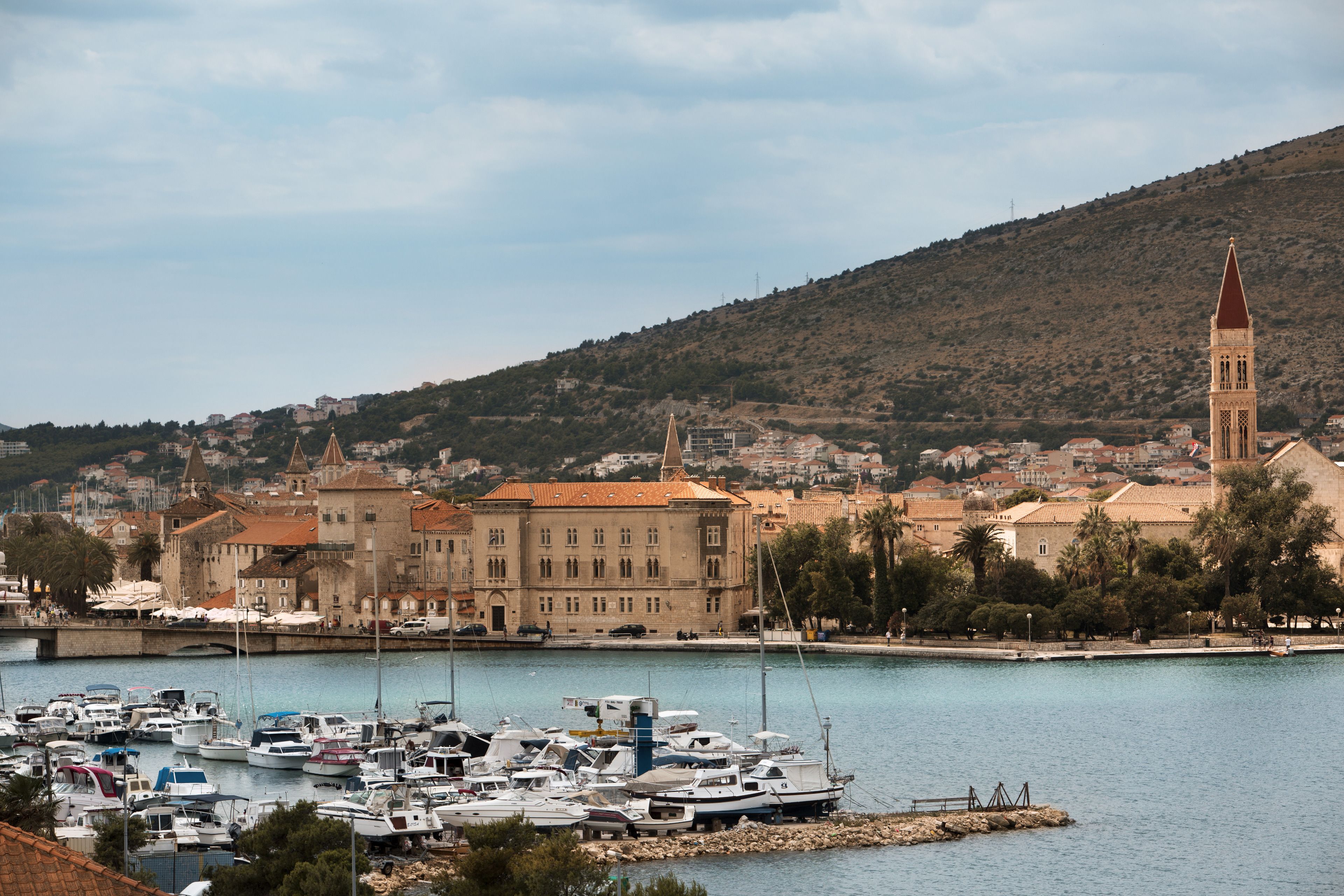 Un port de plaisance avec des bateaux amarrés, une église imposante dotée d’un clocher élevé, et une colline parsemée de bâtiments.