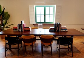 Una mesa de comedor con libros, un vaso y sillas en una habitación con ventana y una planta.
