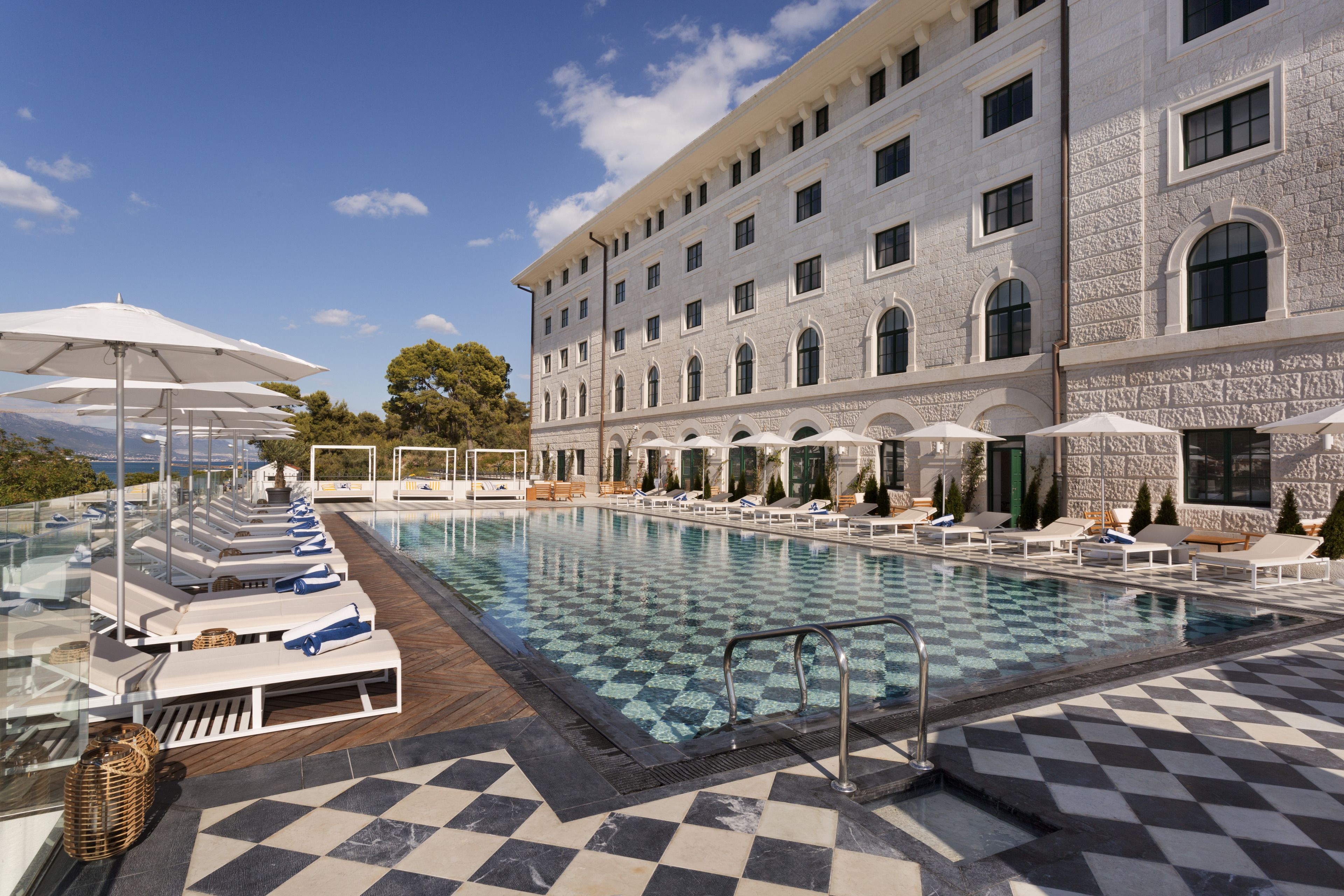 A poolside area with lounge chairs and umbrellas, a checkered floor, and a building with stone facade and arched windows.