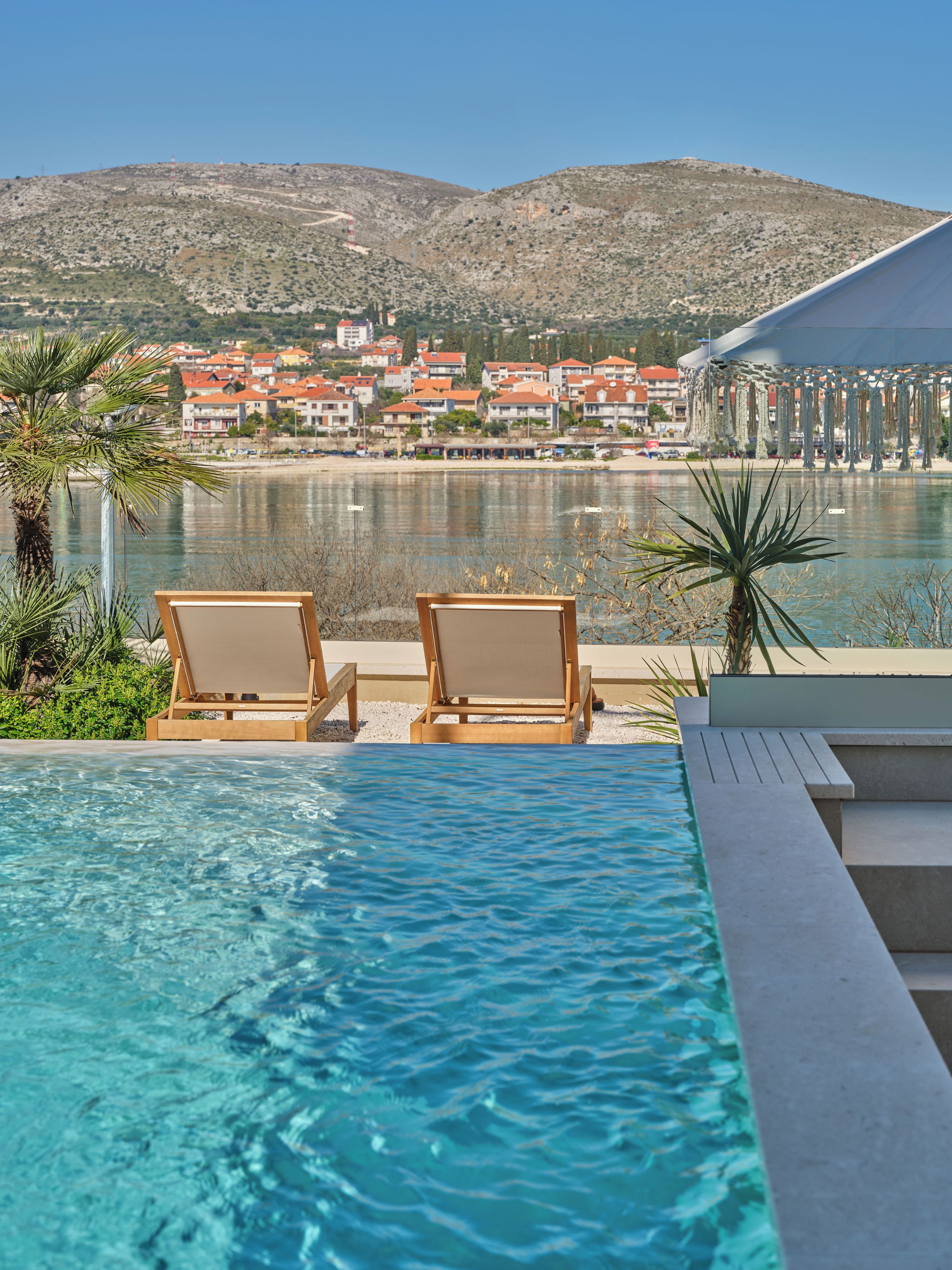 A poolside view with two lounge chairs, a clear view of a body of water, and a town in the background.