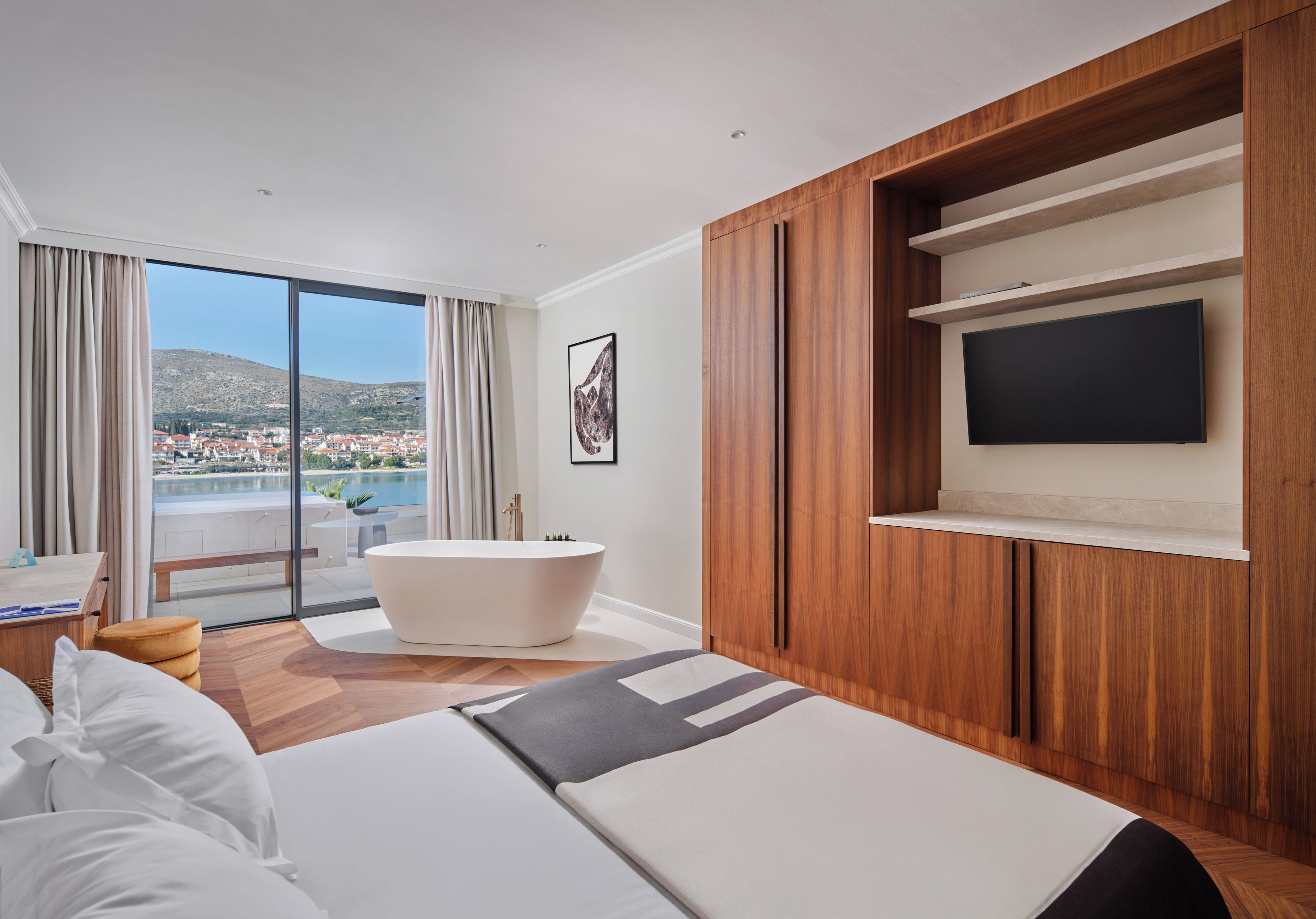 A modern hotel room with a large bed, a bathtub, a TV mounted on the wall, and a view of a cityscape through large windows.