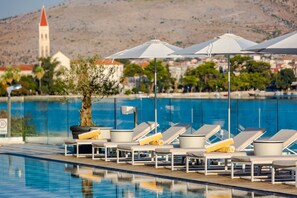 A poolside area with lounge chairs and umbrellas, a tree, and a building with a red roof in the background. - Hotel Brown Beach House & Spa (Trogir)