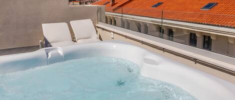 A hot tub on a rooftop with two white lounge chairs and a view of a cityscape and mountains in the background.