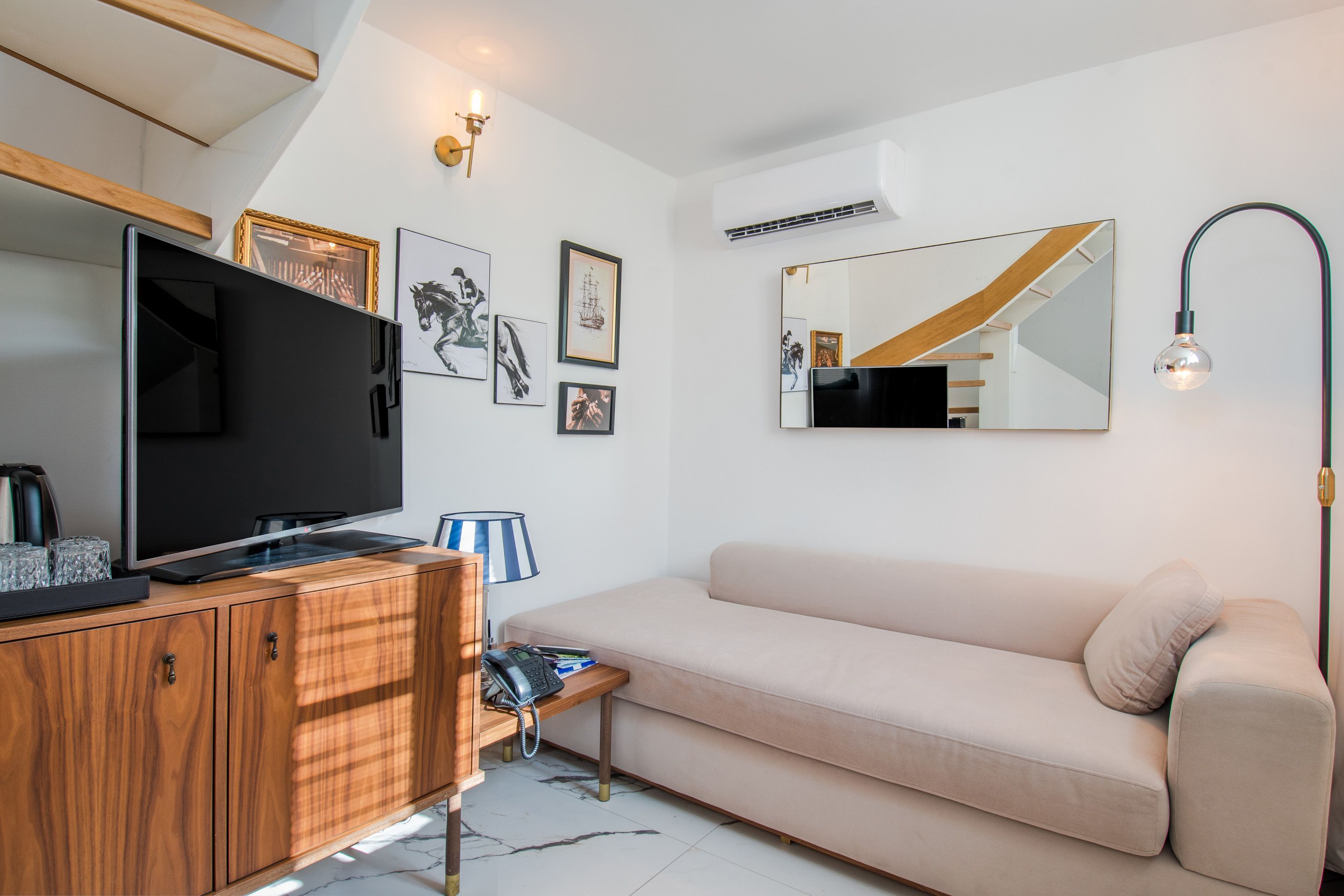 A modern living room with a flat-screen TV, wooden cabinet, beige sofa, and a small table with a lamp.