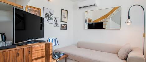 A modern living room with a flat-screen TV, wooden cabinet, beige sofa, and a small table with a lamp.