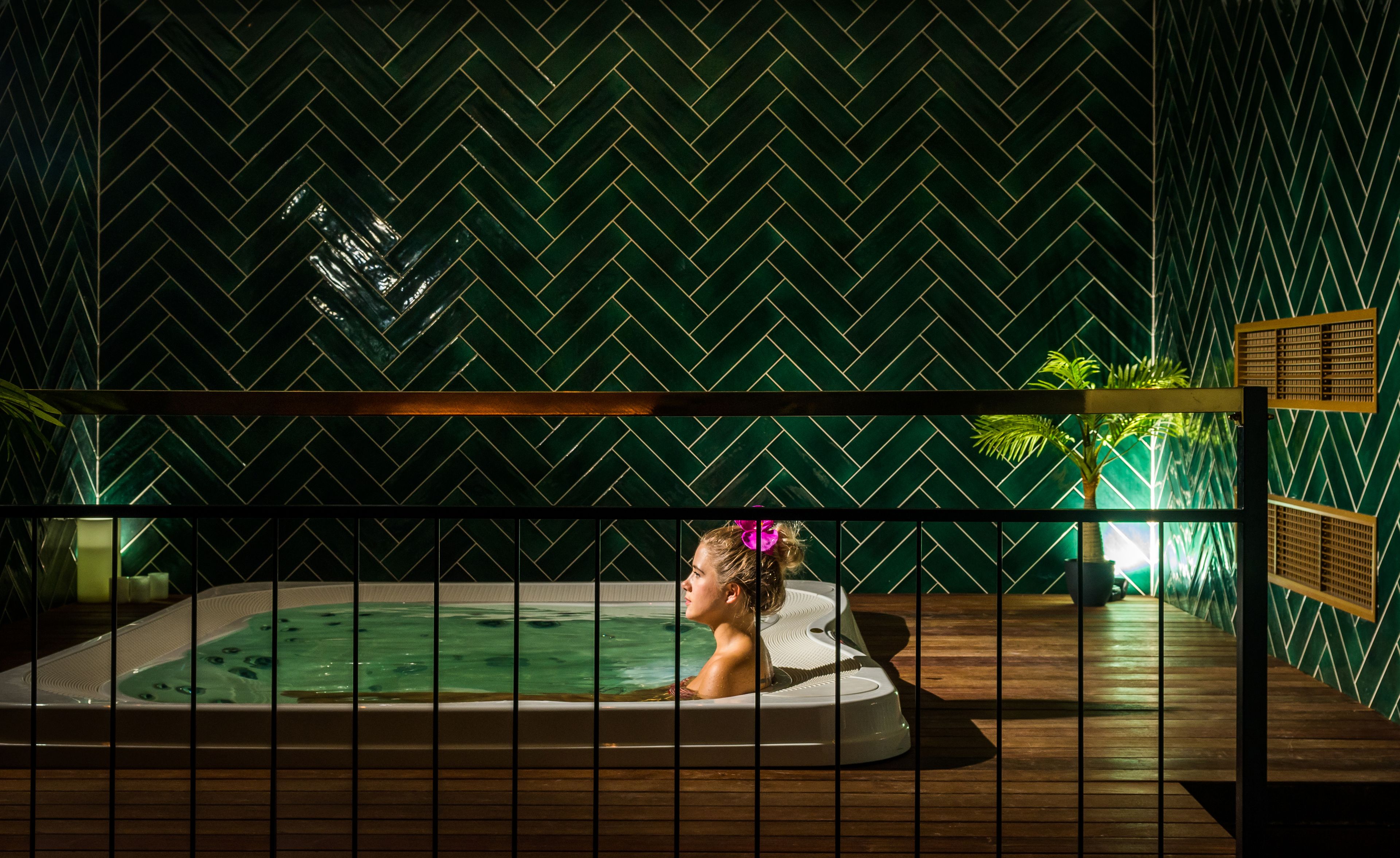 A spa area with a hot tub, wooden deck, and a green herringbone-patterned wall.