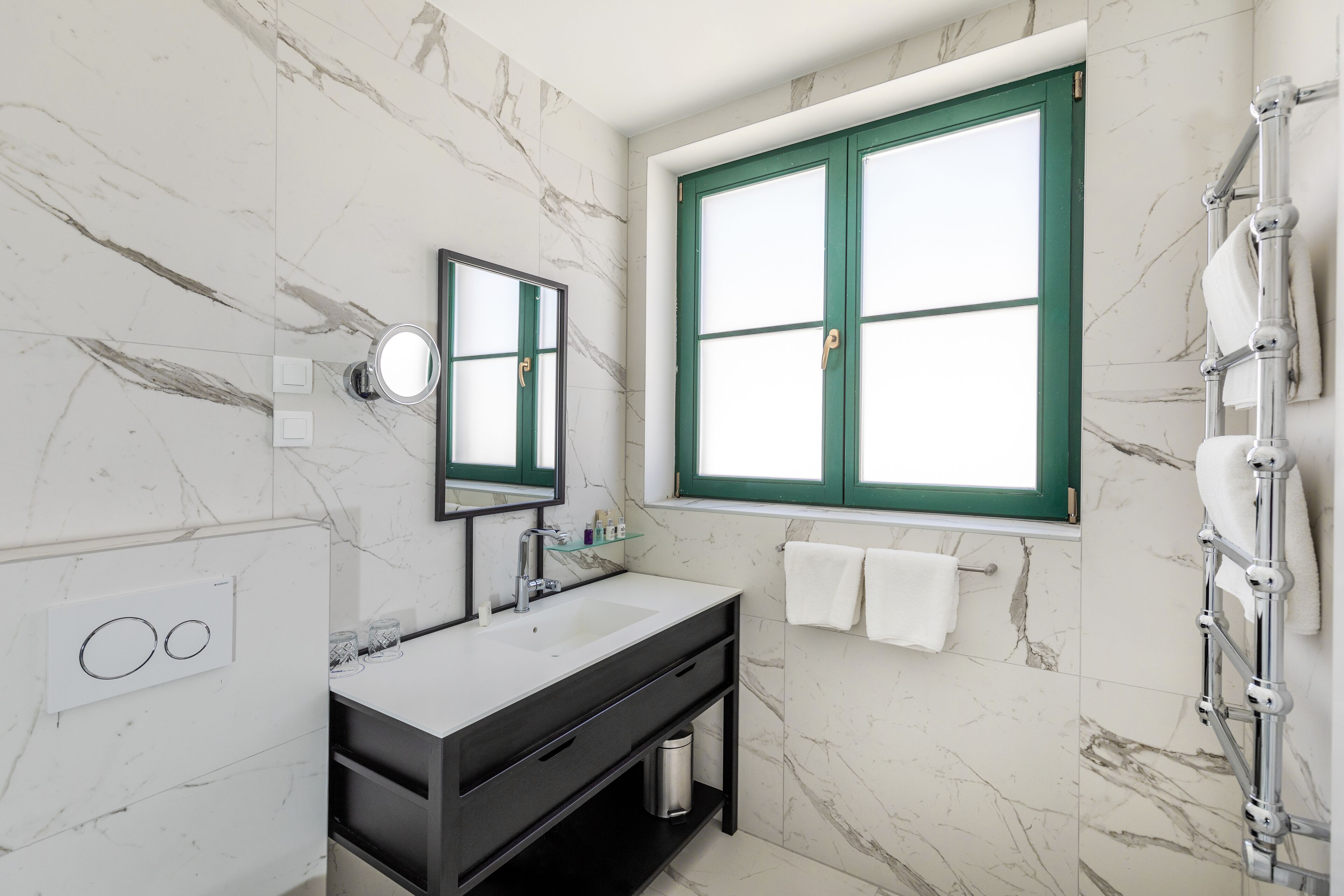 Un bagno con parete in marmo, un lavandino bianco, uno specchio, una finestra con cornice verde e un porta asciugamani.