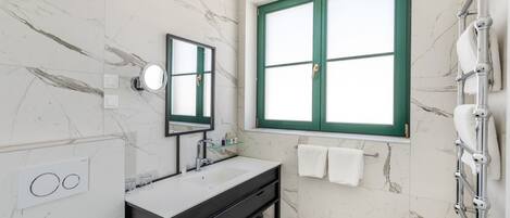 Un bagno con parete in marmo, un lavandino bianco, uno specchio, una finestra con cornice verde e un porta asciugamani.