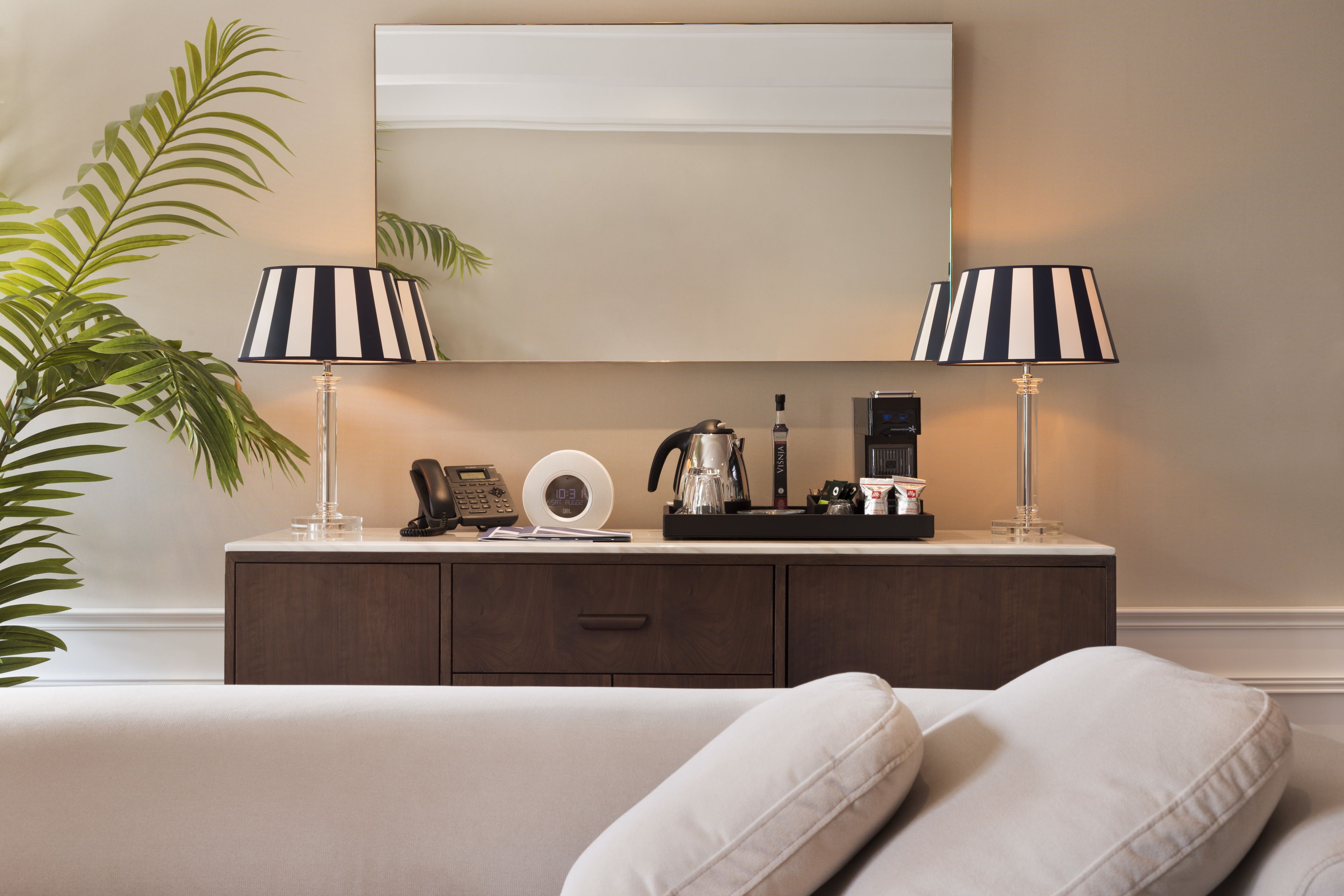 A neatly arranged hotel room with a bed, two bedside lamps, a coffee maker, a clock, and a mirror on the wall.