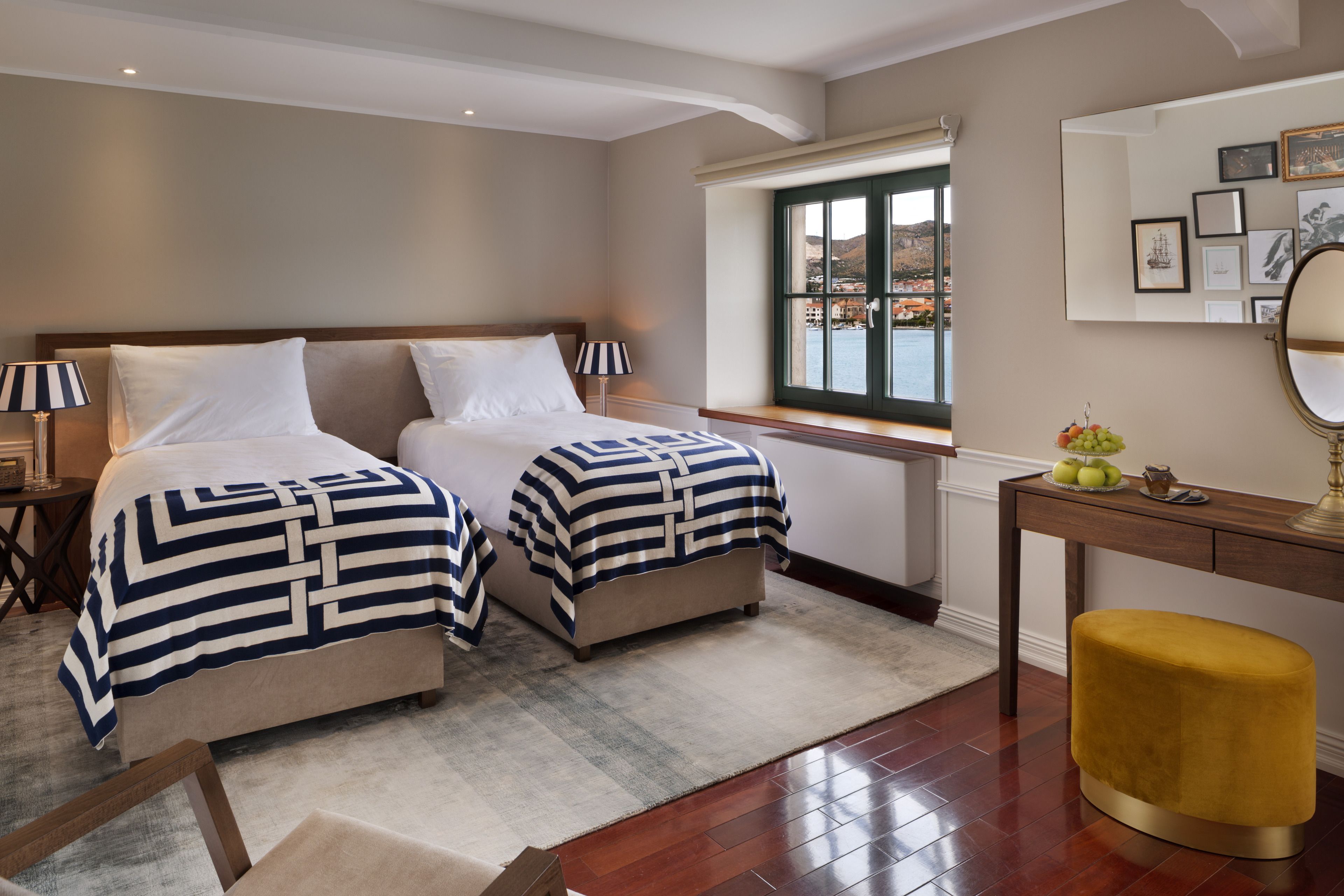Habitación de hotel con dos camas, un escritorio de madera, un reposapiés amarillo, una ventana con vistas y un espejo con fotografías enmarcadas.