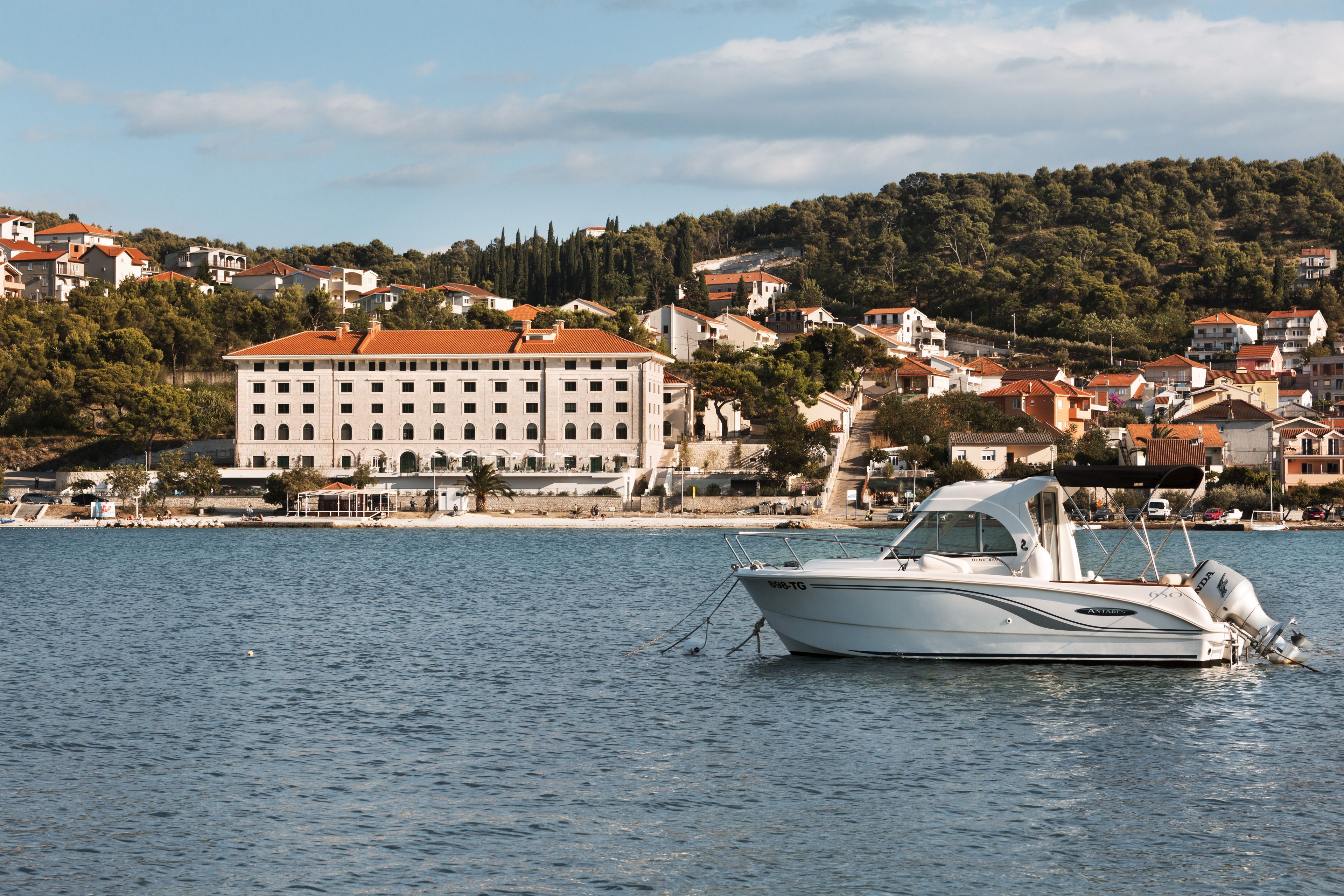 A waterfront property with a large building, a boat docked in the water, and a hillside with residential buildings.
