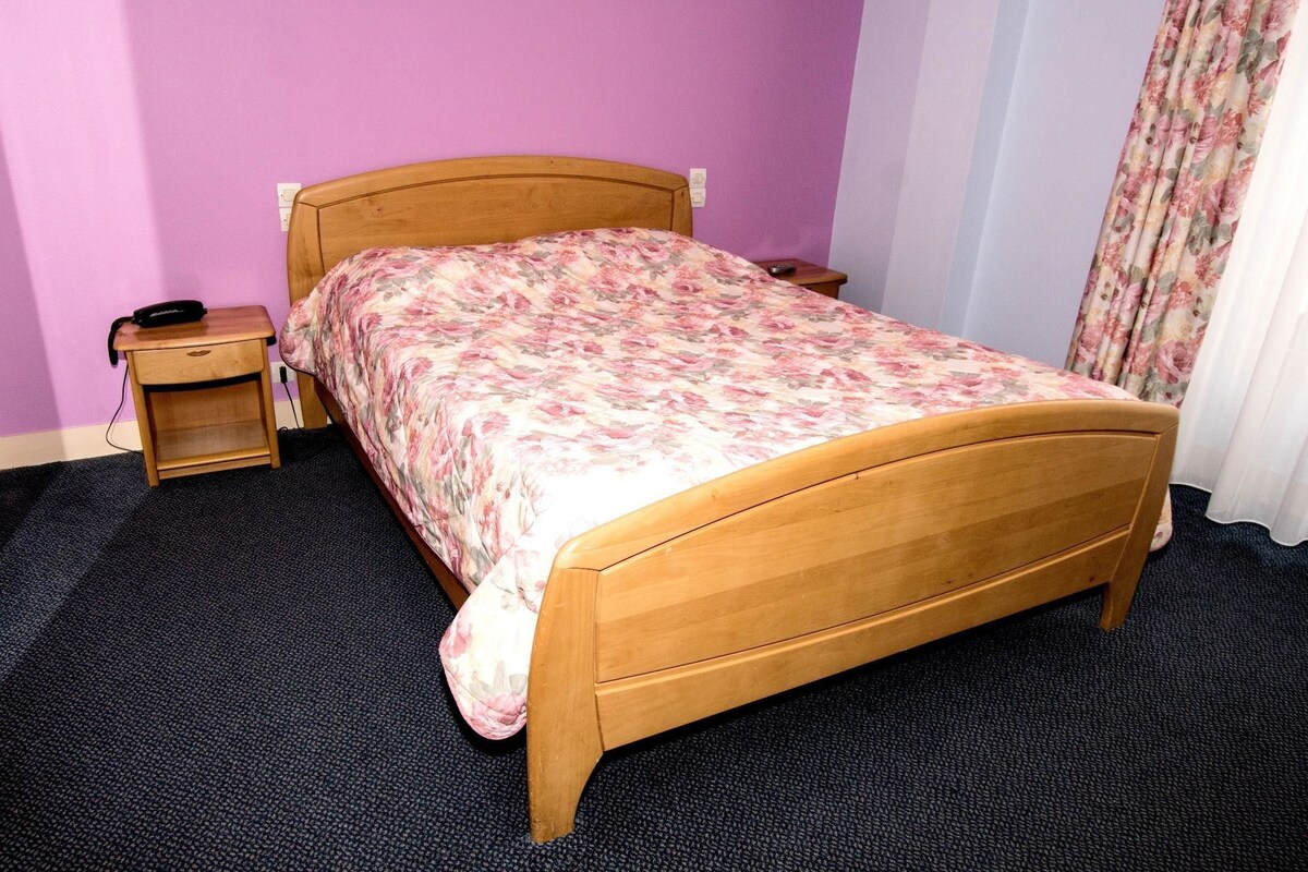Een kamer met een houten bed, een nachtkastje, een wandlamp en een gordijn.