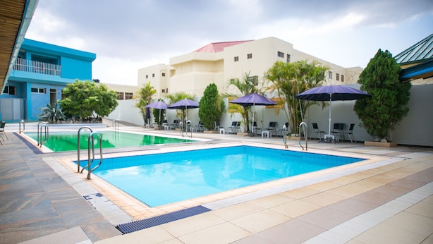 A swimming pool area with two pools, lounge chairs, umbrellas, and a building with a blue and beige facade. - Erata Hotel (Accra)