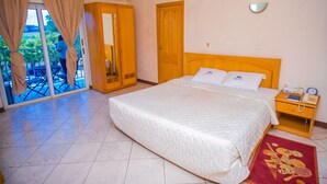 A hotel room with a large bed, a wooden nightstand, a telephone, and a view through a glass door to an outdoor area with a table and chairs. - Erata Hotel (Accra)