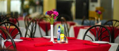 Comedor con mesas dispuestas, manteles rojos, sillas de metal y un jarrón con flores.