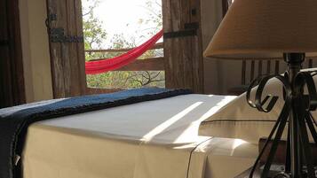 A room with a bed covered in a beige sheet, a lamp on a tripod, and a view of a red fabric outside a wooden door.