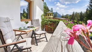 A patio with two cushioned chairs and a wooden table, a window overlooking a mountain, and a wooden deck with flowers. - Das Habitat (Leutasch)
