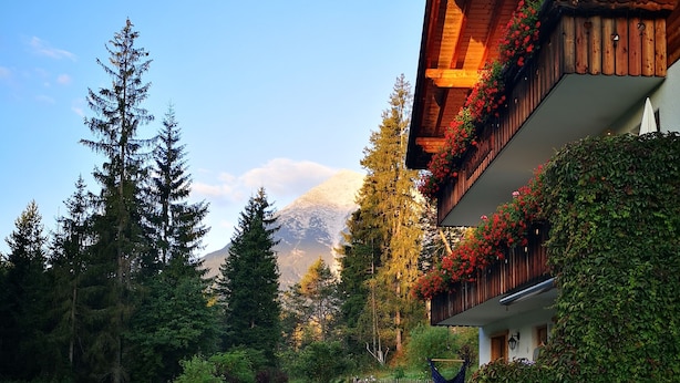 A building with a wooden balcony adorned with flowers, surrounded by a lush garden and a mountain in the background. - Das Habitat (Leutasch)