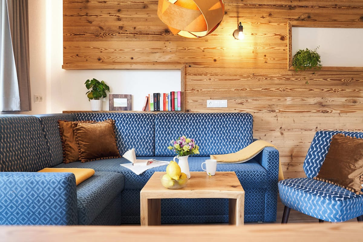 A modern living room with a blue patterned sofa, a wooden coffee table with a vase of flowers, and a bookshelf with books and a small plant.