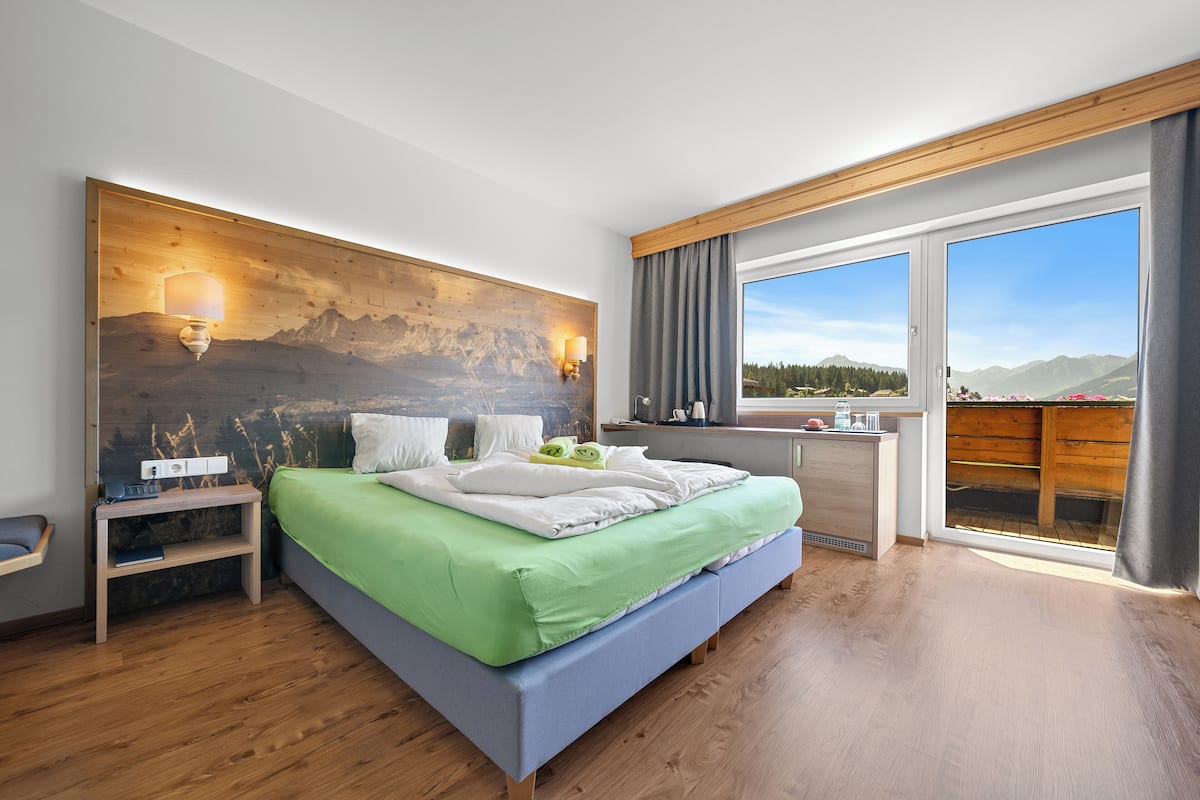 A modern bedroom with a large bed, a wooden headboard, a nightstand, a desk, and a view of mountains through the window.