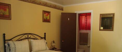 Una cama individual con una manta azul, una mesita de noche con una lámpara, una puerta a un baño y dos cuadros en la pared.