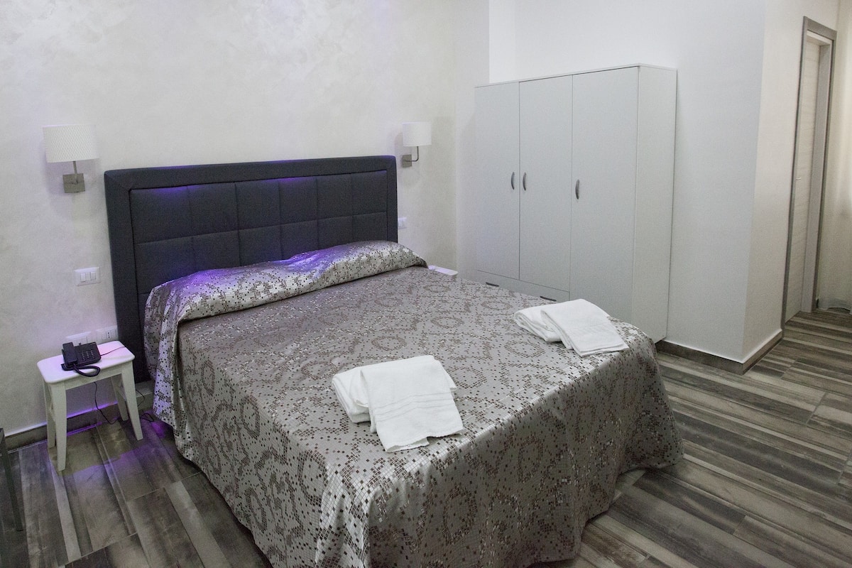 A bedroom with a bed, a nightstand with a phone, a wardrobe, and a mirror.