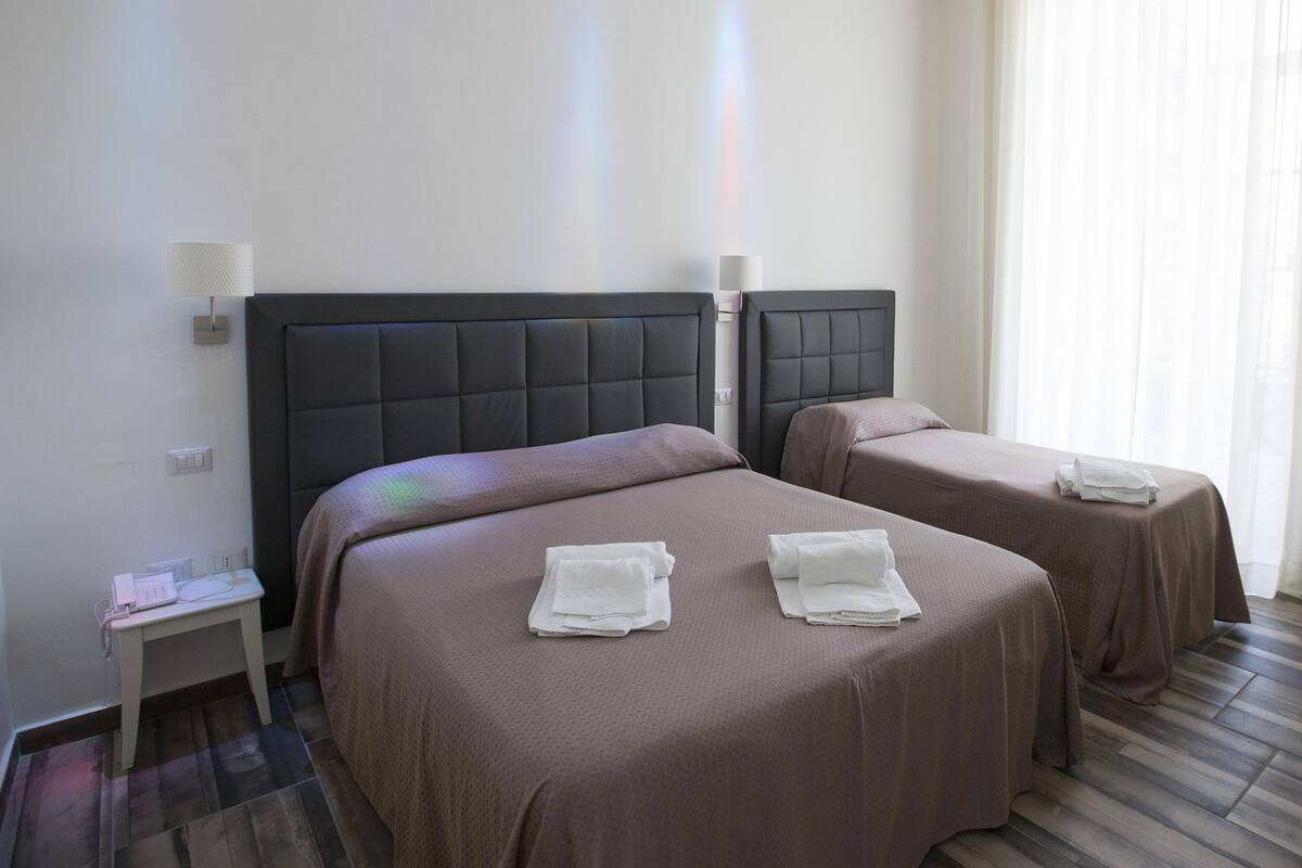 A hotel room with two beds, each with a brown bedspread and white towels. There is a small bedside table with a lamp, a phone, and a power outlet. The room has a wooden floor and a window with sheer curtains.