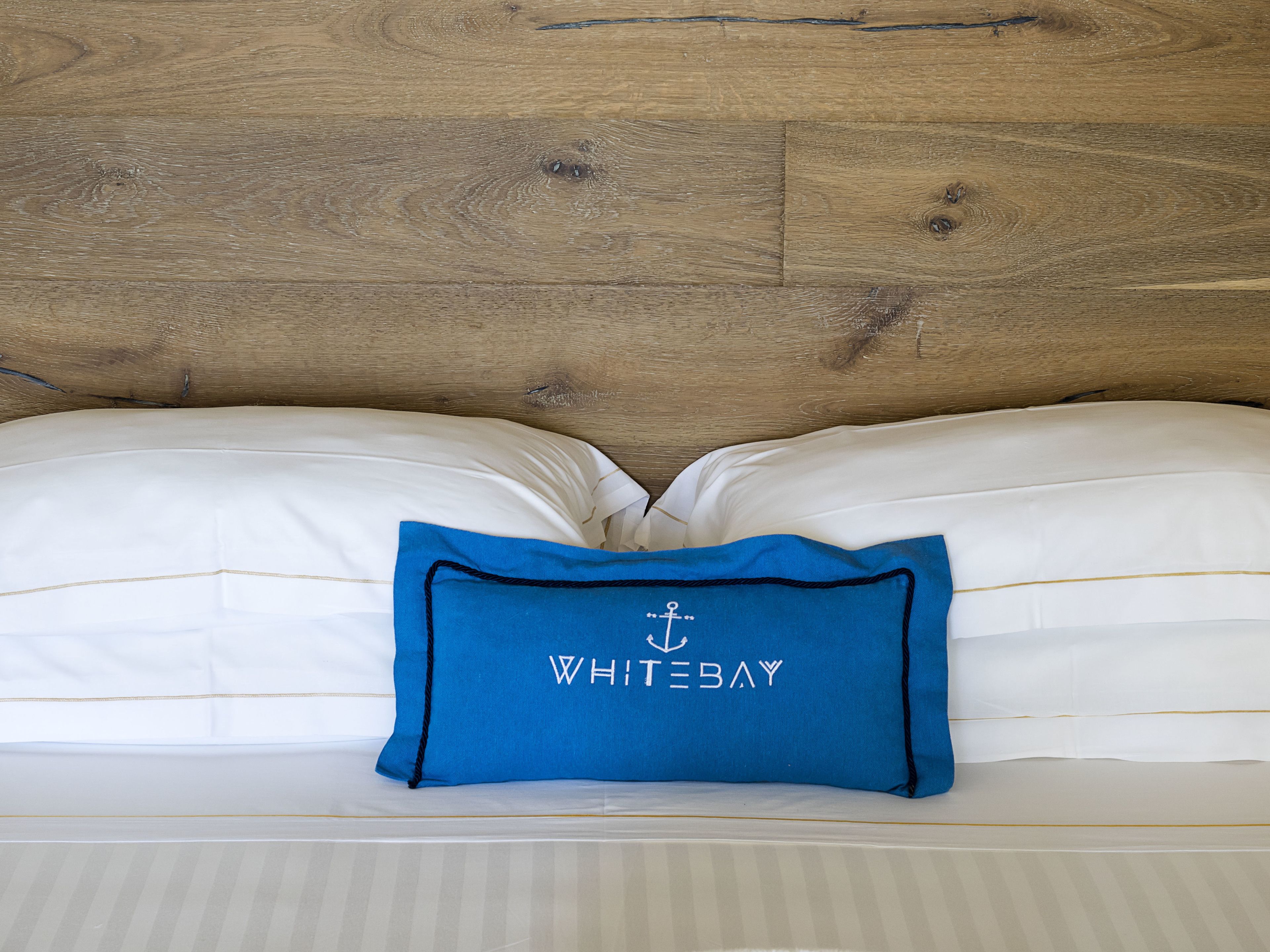 A bed with white linens and a blue pillow with "WHITEBAY" embroidered on it.