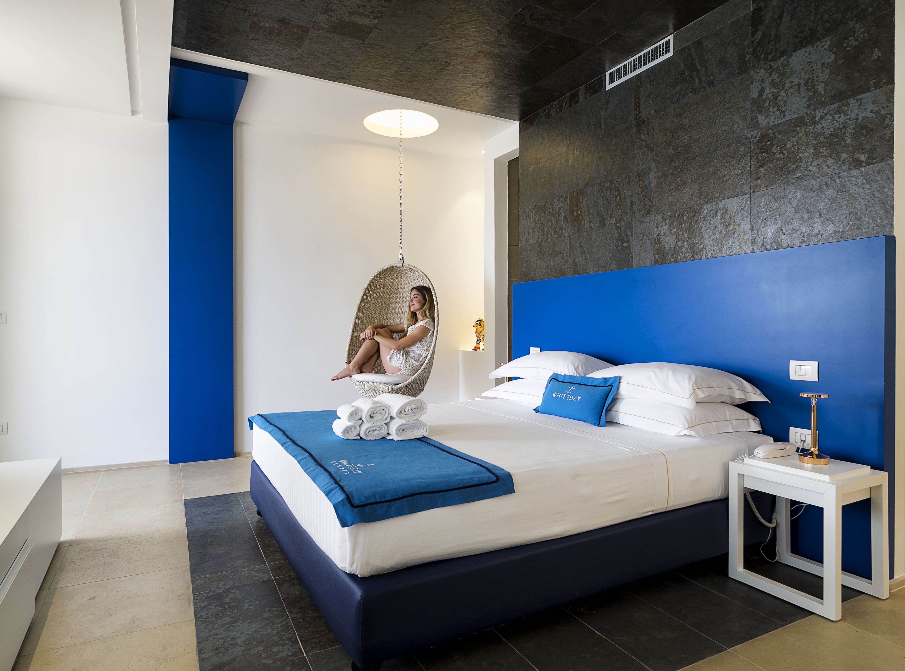 A modern bedroom with a hanging chair, a neatly made bed with blue bedding, and a blue accent wall.