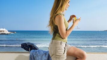 Eine Frau sitzt am Meer, hält ein Getränk und hat Handtuch und Jacke in der Nähe.