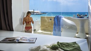 A room with a large window overlooking the sea, a bathtub, a table with a magazine, and a towel hanging on the wall. - White Bay Resort (Syracuse)