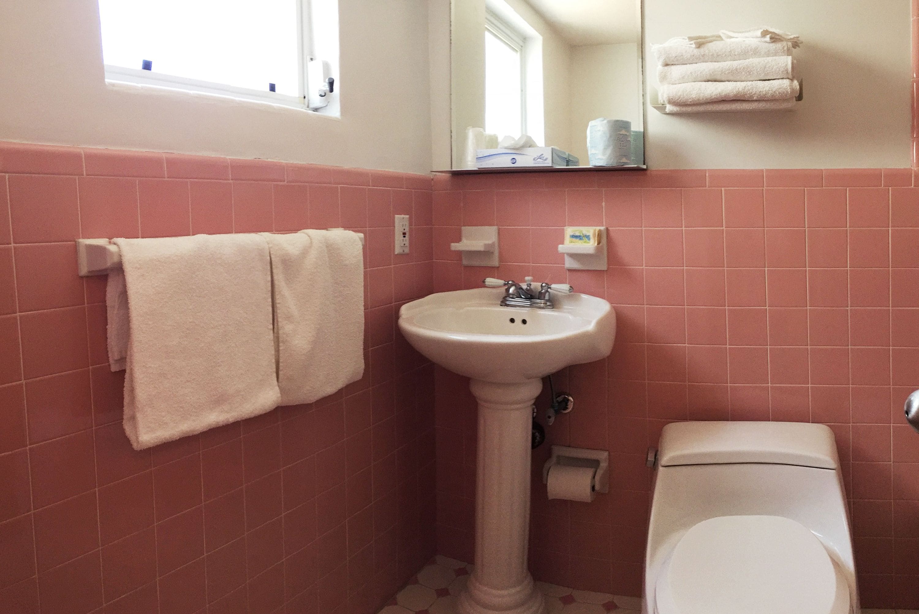 A bathroom with pink tiles, a pedestal sink, a toilet, and a window.