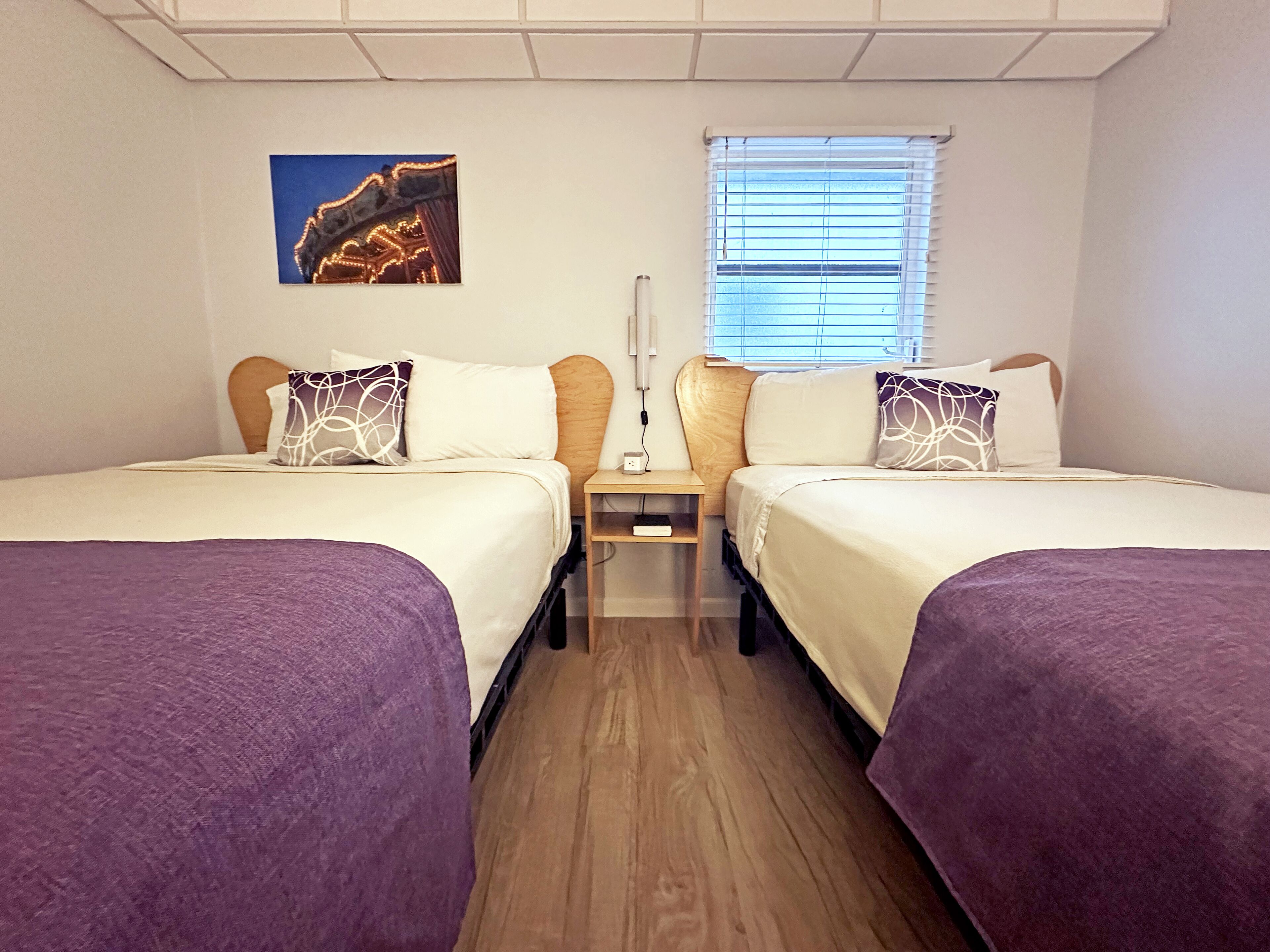 A hotel room with two beds, a nightstand, a lamp, a window with blinds, and a picture on the wall.