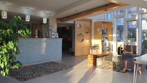 A modern hotel lobby with a reception desk, a seating area with a patterned rug, and large windows offering a view of the outside. - Shalimar Resort (Wildwood Crest)