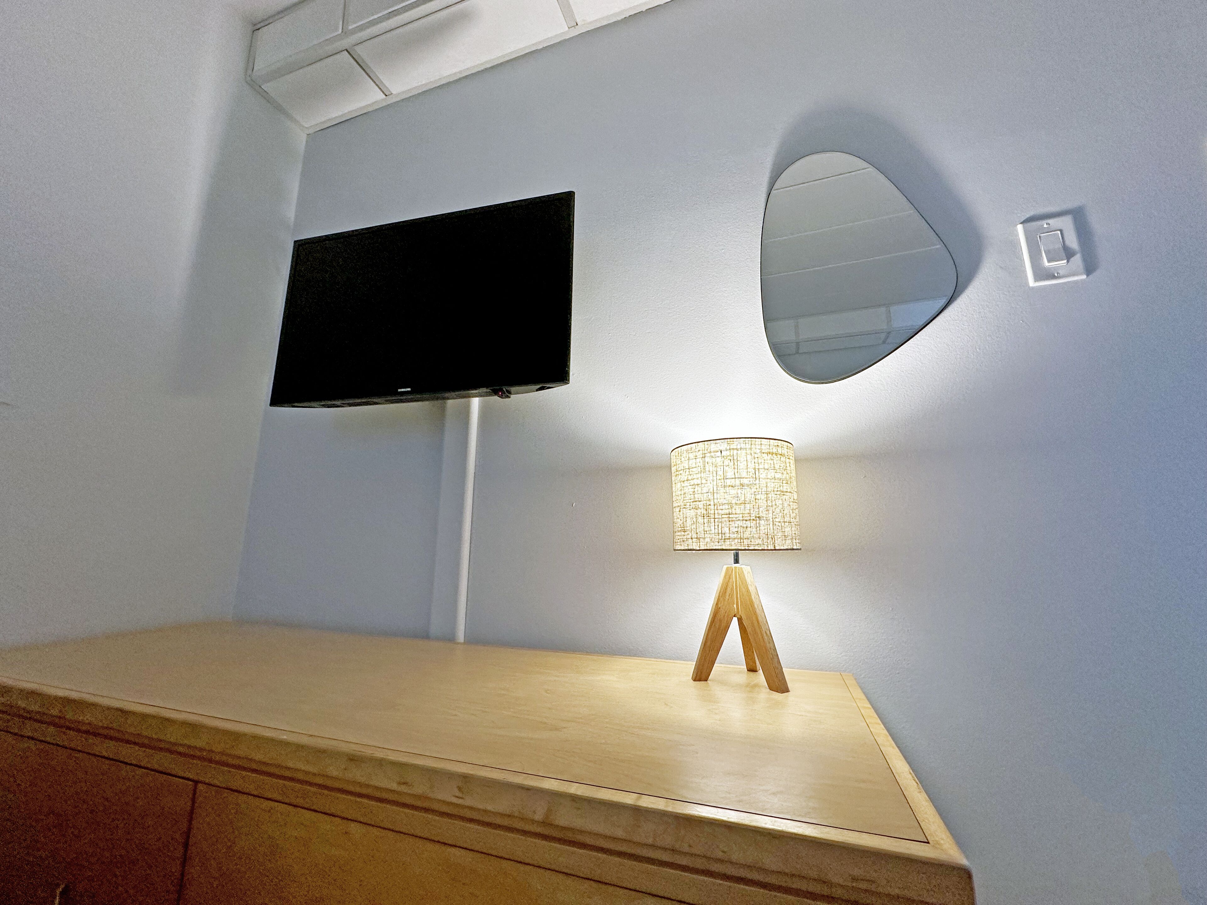 A room with a flat-screen TV mounted on the wall, a round mirror, and a wooden table with a lamp.