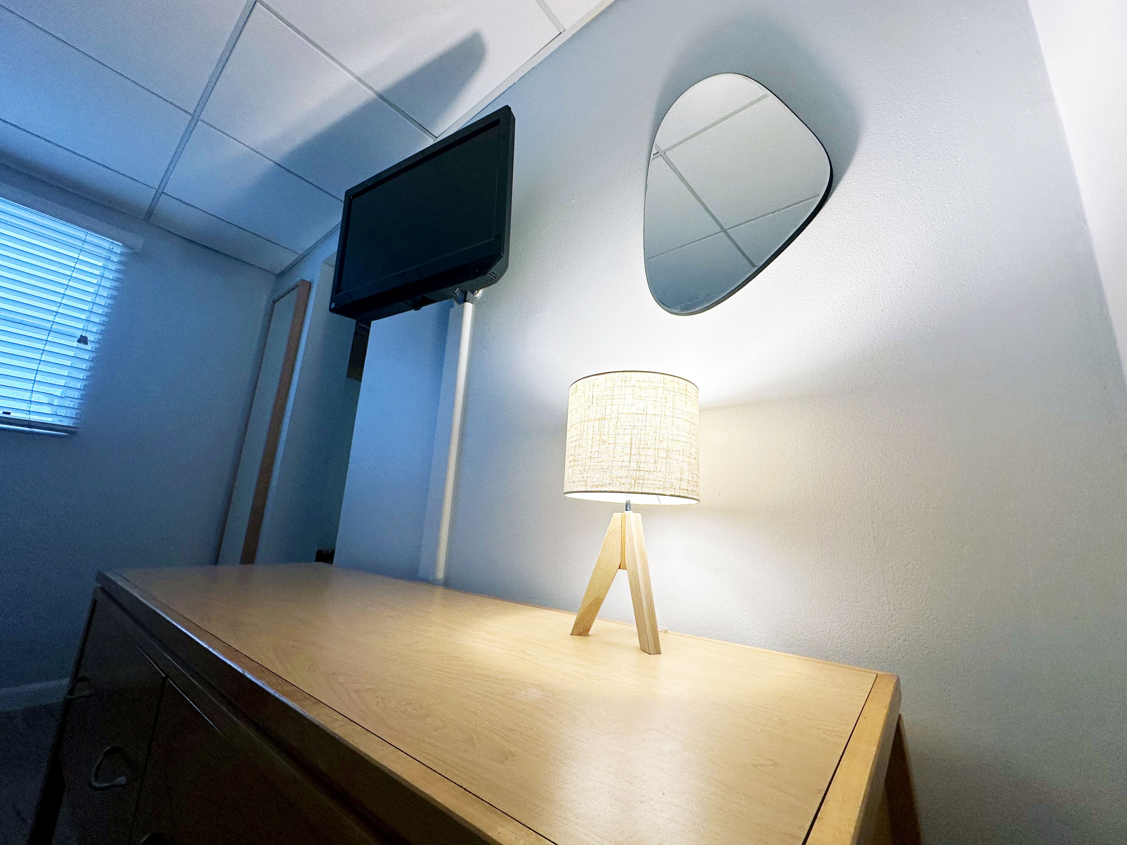 Una habitación con un escritorio de madera, un televisor en la pared, una lámpara y un espejo.