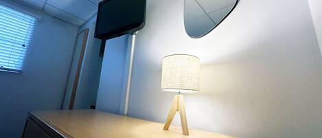 Una habitación con un escritorio de madera, un televisor en la pared, una lámpara y un espejo.