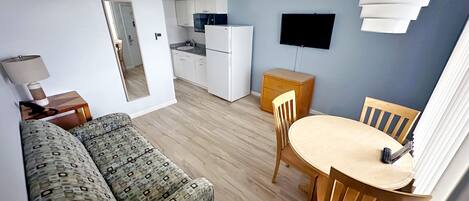 A compact living space with a sofa, a dining table with chairs, a small kitchenette, and a TV mounted on the wall.