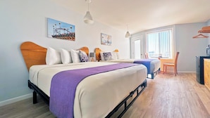 A modern bedroom with a large bed, a smaller bed, a desk, and a chair. There are two windows with blinds, a hanging light fixture, and artwork on the walls. - Shalimar Resort (Wildwood Crest)