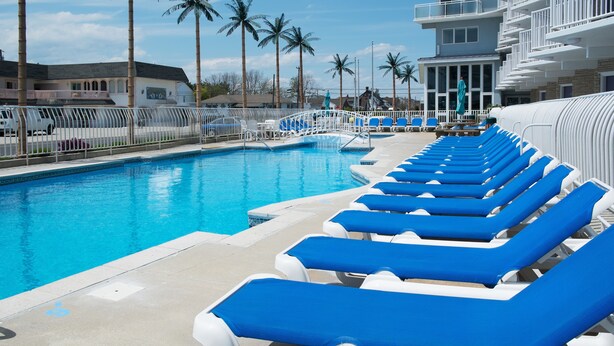 A swimming pool with blue lounge chairs, palm trees, and a building with balconies. - Shalimar Resort (Wildwood Crest)
