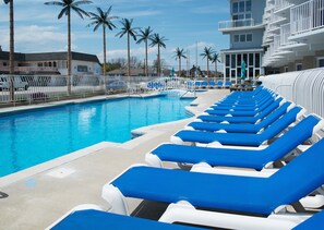 Una piscina con sillas reclinables azules, palmeras y un edificio con balcones.