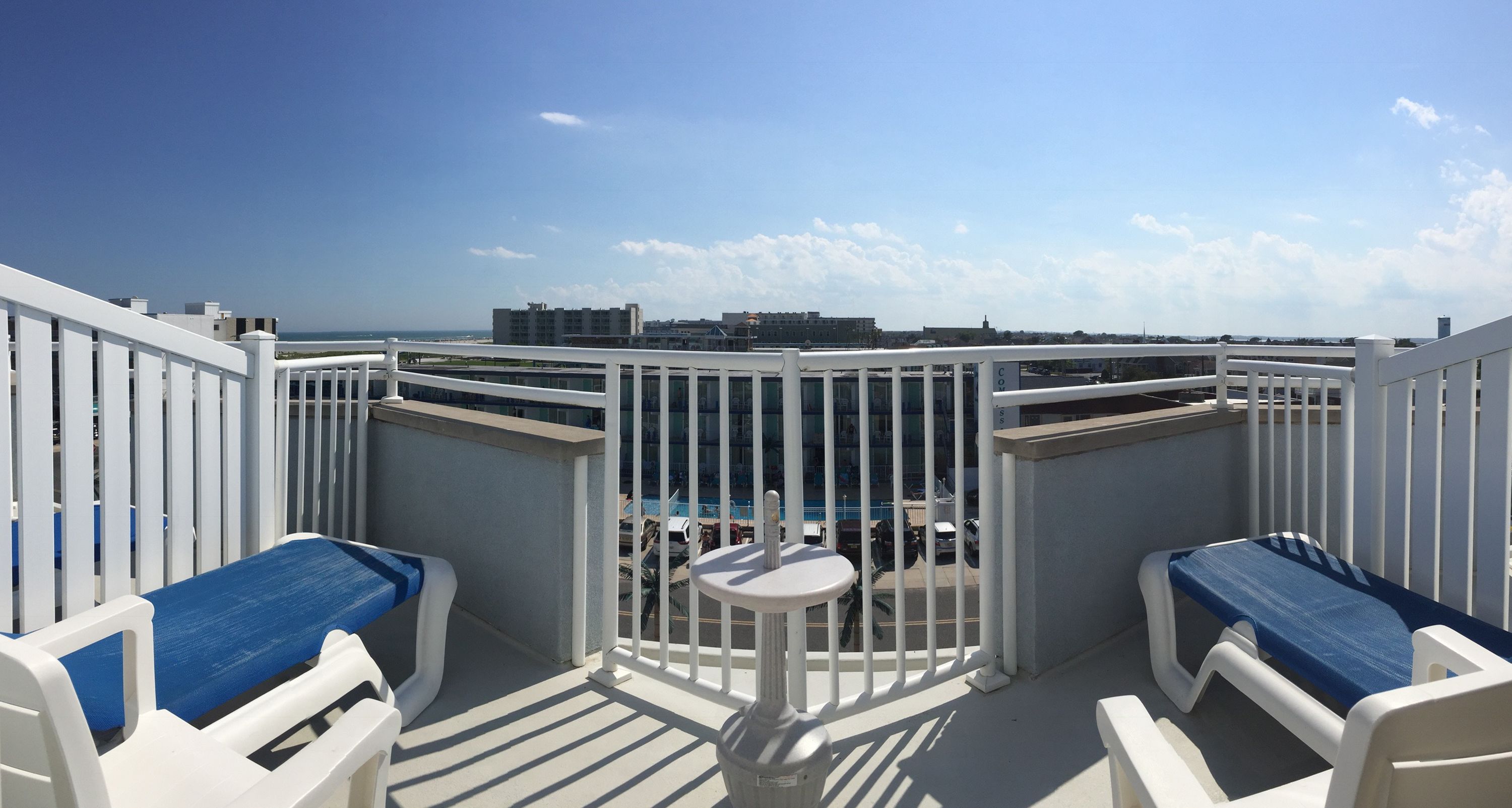 Ein Balkon mit weißen Geländern, zwei blaue Sitzgelegenheiten mit Kissen, einem kleinen runden Tisch und Blick auf Stadt und Meer.